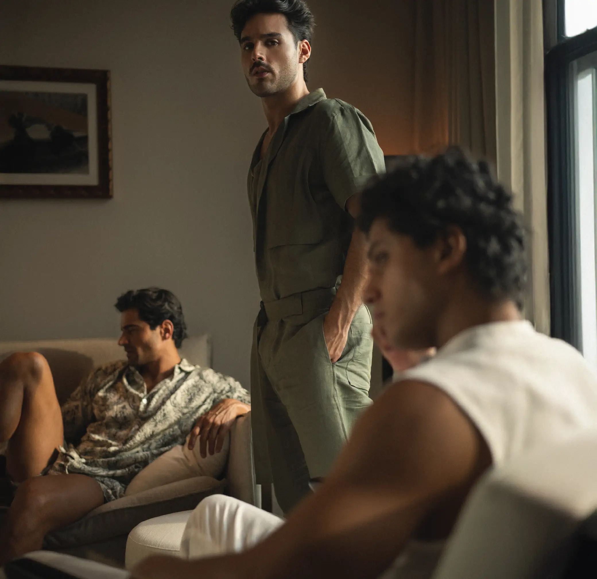 Three men in a warm, softly lit room. One stands in a green suit, another lounges in a patterned outfit, and the third sits slightly out of focus in the foreground – calm and introspective