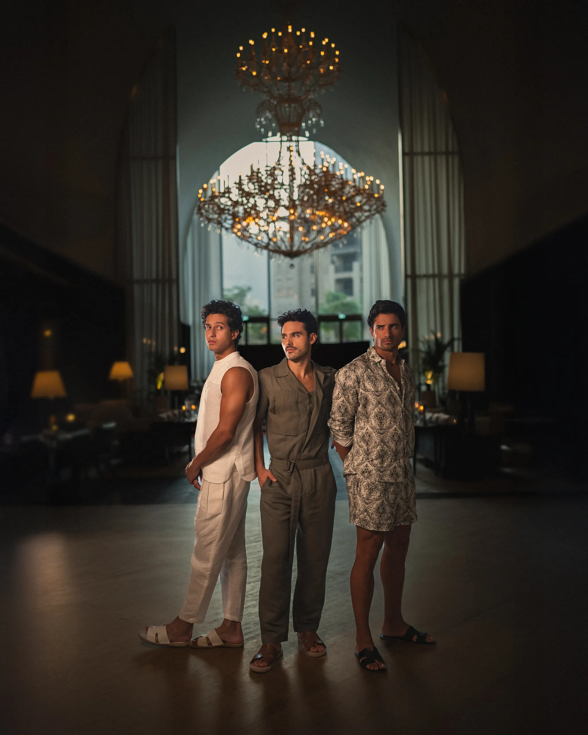 Three men in stylish 20Two Elemental garments standing beneath a grand chandelier in a dimly lit, luxurious interior with arched windows and ambient lighting and upscale decor