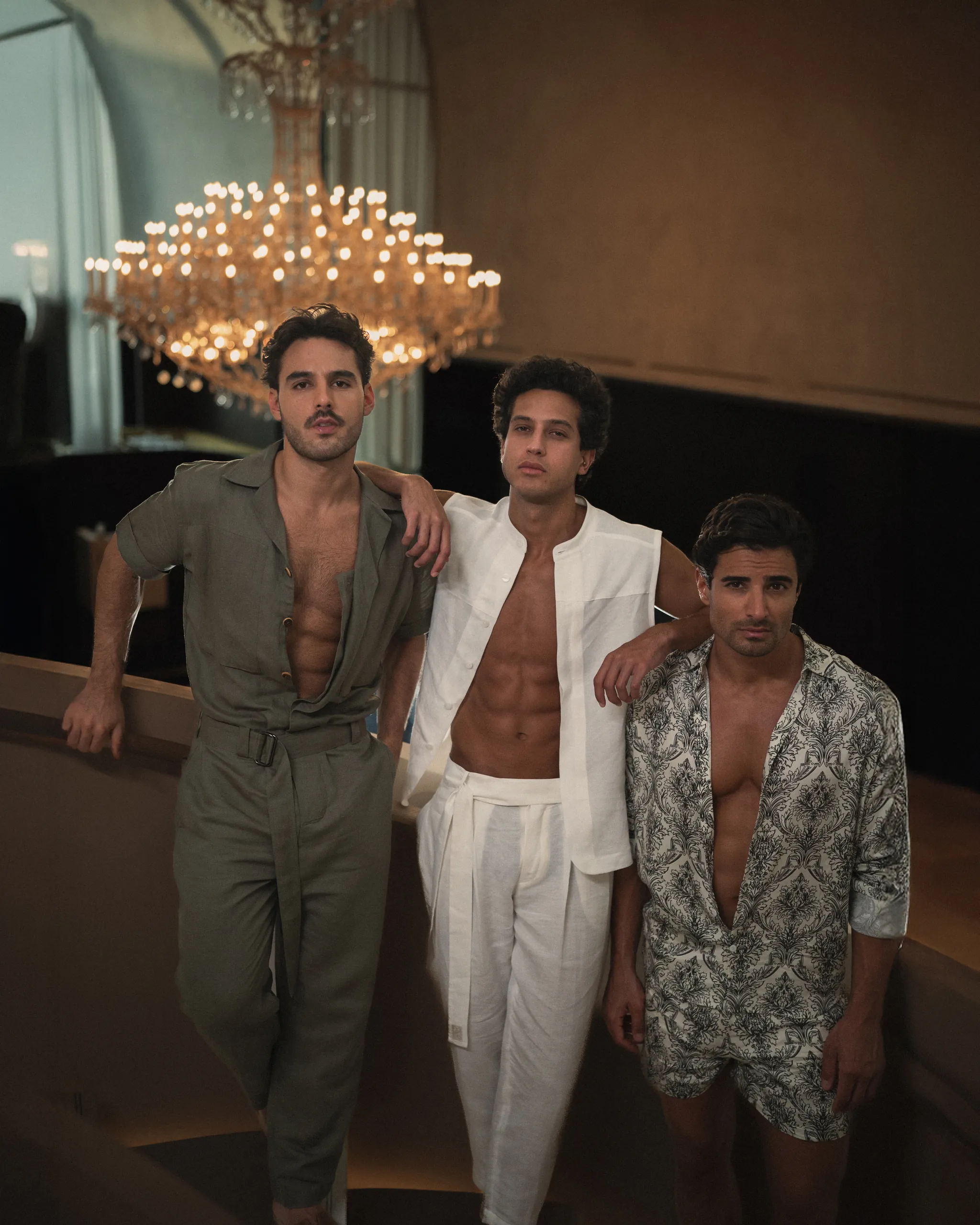 Three men in stylish, 20Two Elemental outfits standing beneath an ornate chandelier in an elegant indoor setting. Warm lighting and upscale decor frame the scene, blending modern fashion with a luxurious atmosphere