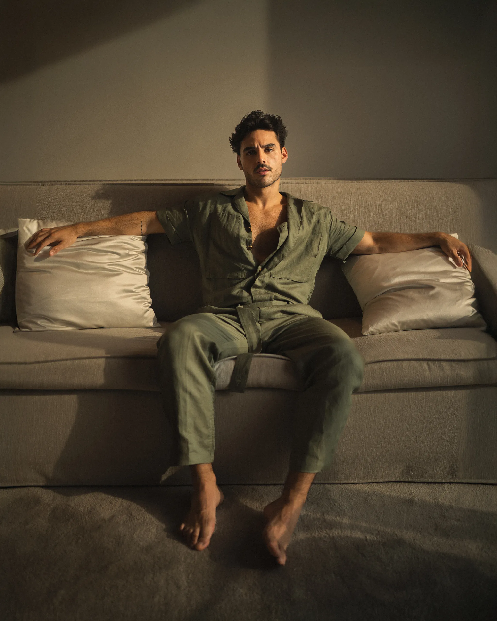 Relaxed man in a green 20Two Elemental suit with arms stretched along the back of a beige couch, framed by soft pillows and dramatic lighting, creating a moody, editorial atmosphere