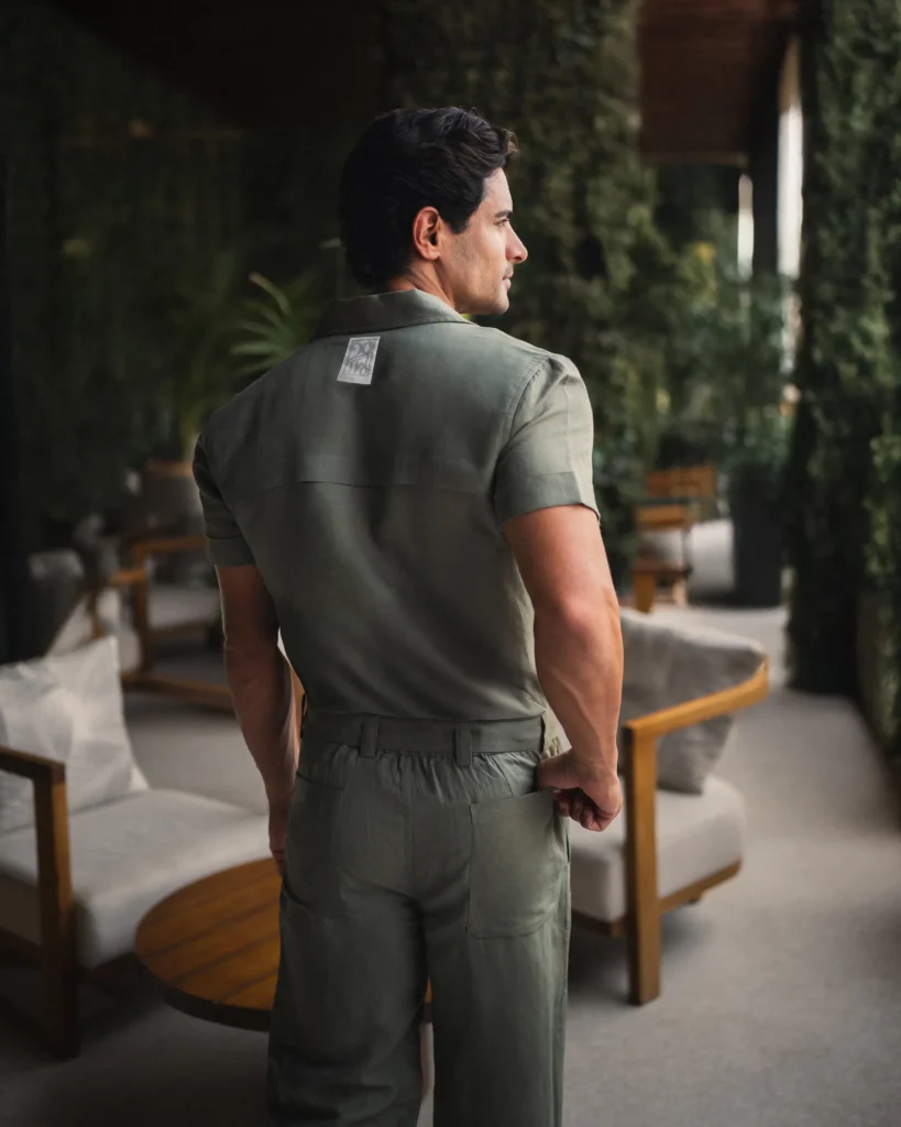 Man in a green 20Two Elemental suite with a Cuban collar, standing in a serene outdoor lounge surrounded by tall greenery. Brown leather sandals and breathable fabric suggest a relaxed vibe, with cushioned seating and a wooden table adding to the tranquil, design-forward atmosphere, back view
