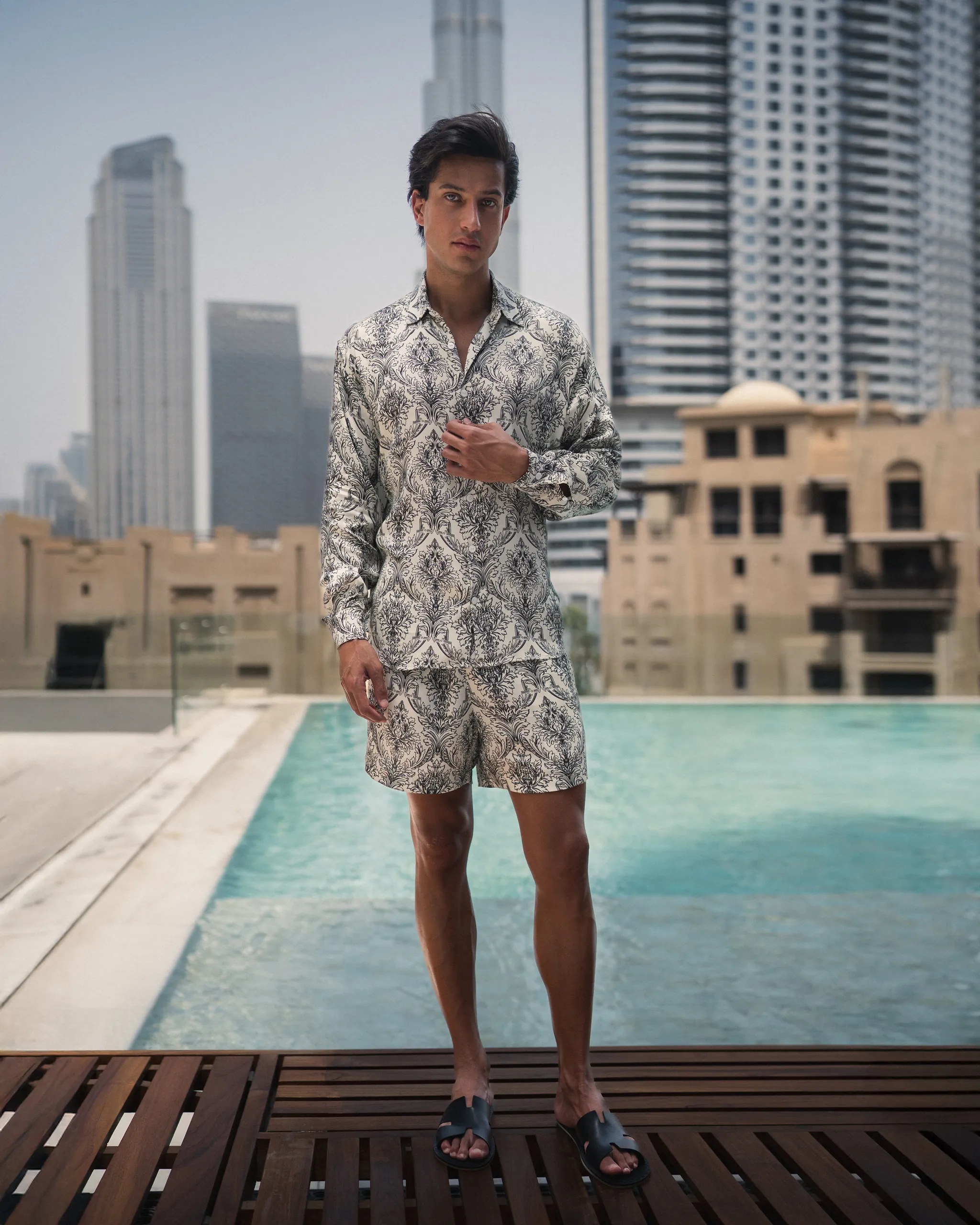 Man wearing a refined and styled patterned lotus plant fiber shirt and matching shorts from 20Two Elemental, standing by a pool with a modern city skyline in the background, front view