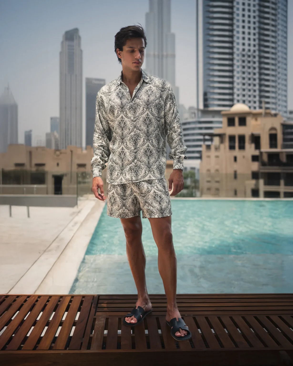 Man wearing a refined and styled patterned lotus plant fiber shirt and matching shorts from 20Two Elemental, standing by a pool with a modern city skyline in the background, front view