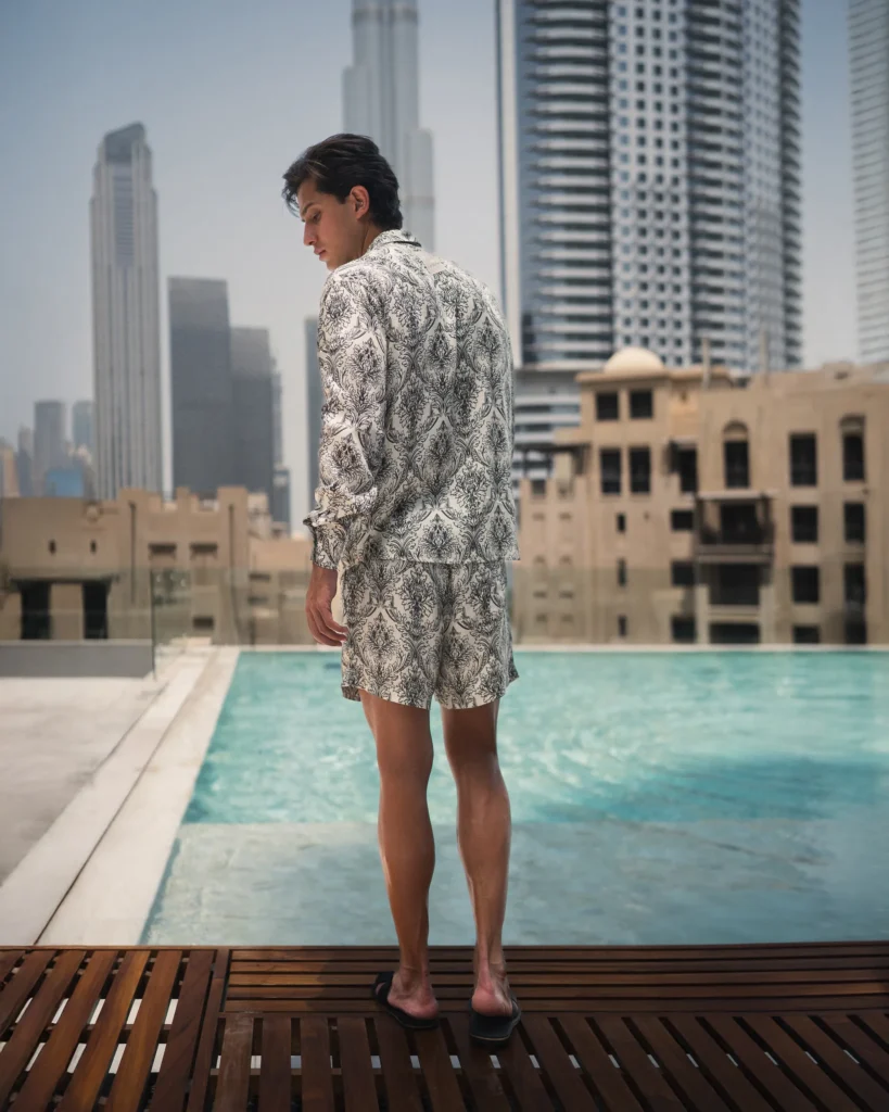 Man wearing a refined and styled patterned lotus plant fiber shirt and matching shorts from 20Two Elemental, standing by a pool with a modern city skyline in the background, back view