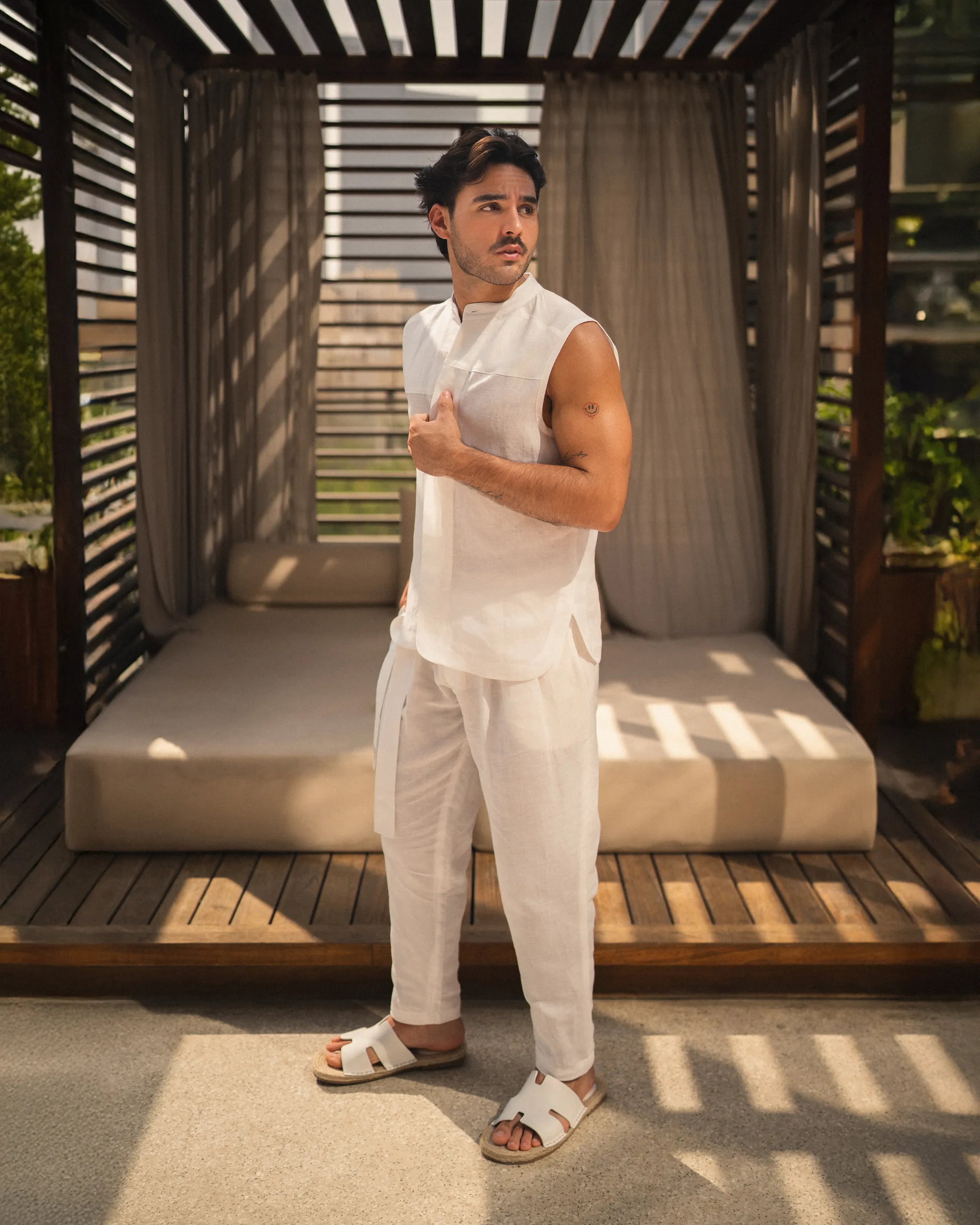 Confident man wearing a stylish natural white, hemp and bamboo blend sleeveless shirt with mandarin collar and long pant set from 20Two Elemental, standing in front of a modern outdoor cabana with filtered sunlight, front view