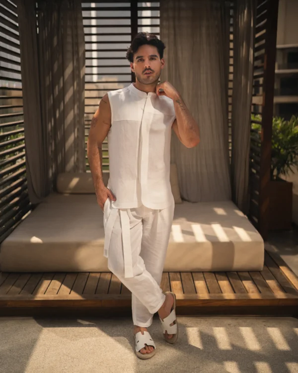 Confident man wearing a stylish natural white, hemp and bamboo blend sleeveless shirt with mandarin collar and long pant set from 20Two Elemental, standing in front of a modern outdoor cabana with filtered sunlight, front view