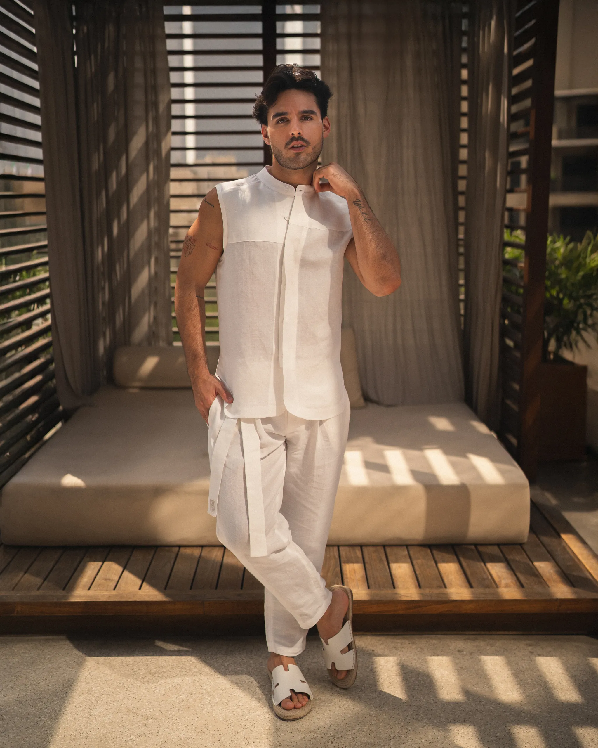 Confident man wearing a stylish natural white, hemp and bamboo blend sleeveless shirt with mandarin collar and long pant set from 20Two Elemental, standing in front of a modern outdoor cabana with filtered sunlight, front view
