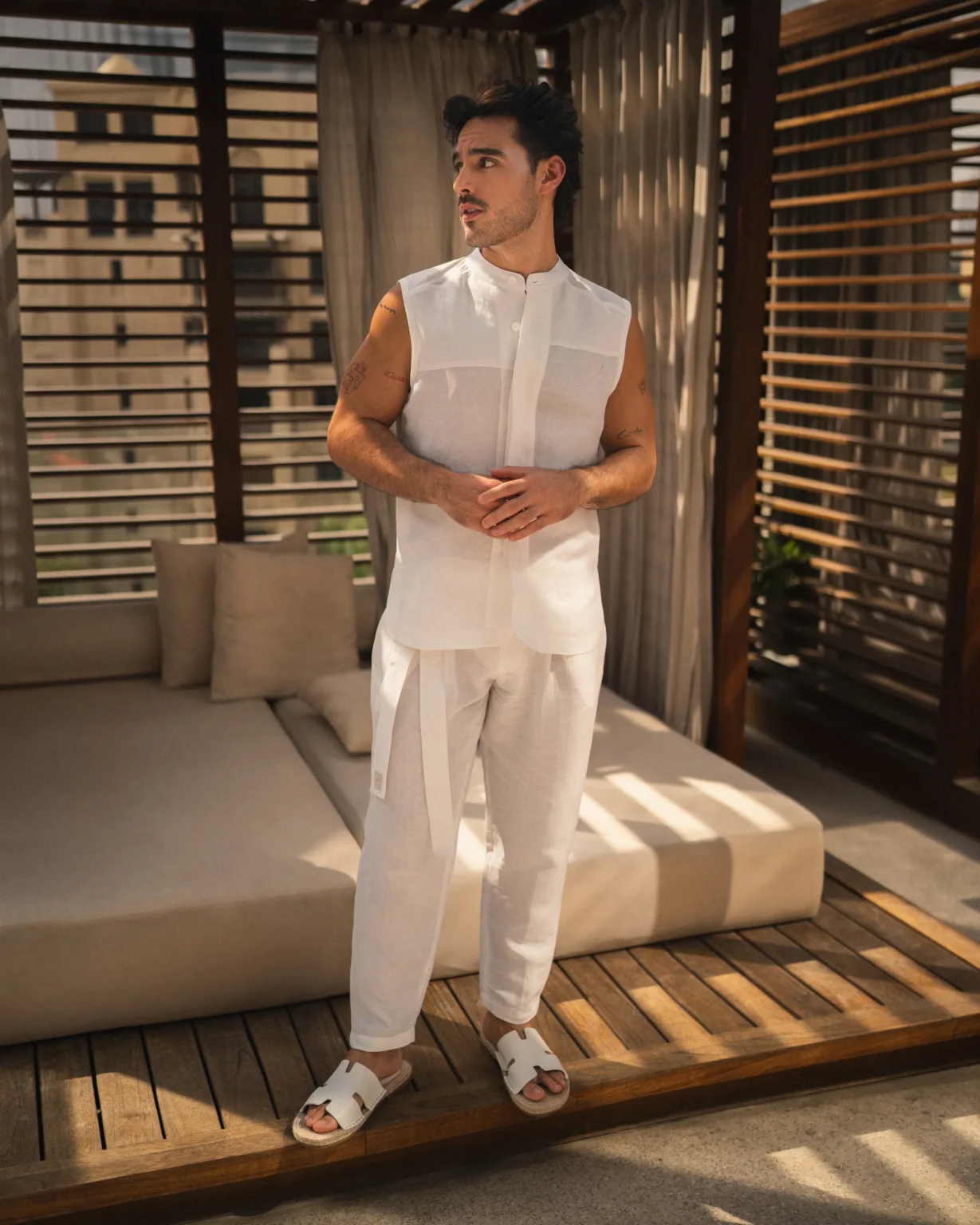 Confident man wearing a stylish natural white, hemp and bamboo blend sleeveless shirt with mandarin collar and long pant set from 20Two Elemental, standing in front of a modern outdoor cabana with filtered sunlight, front view
